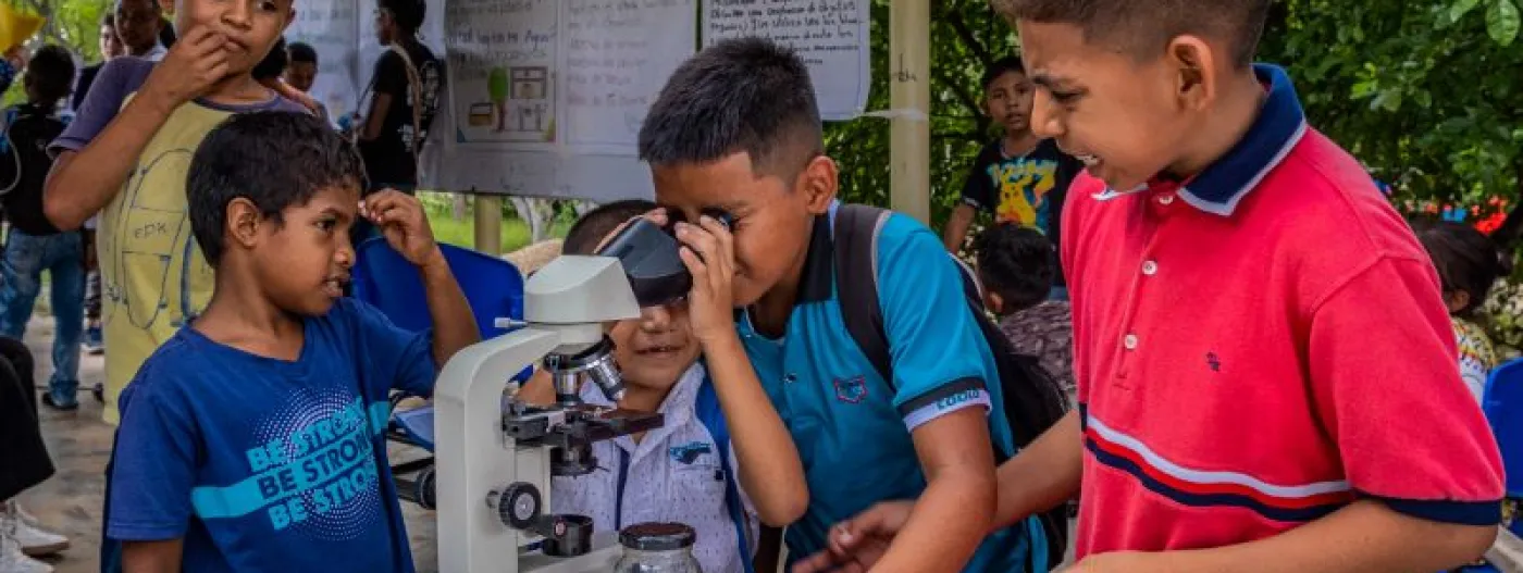 Grupo de cinco niños junto a un microscopio y uno de ellos mira a través del lente