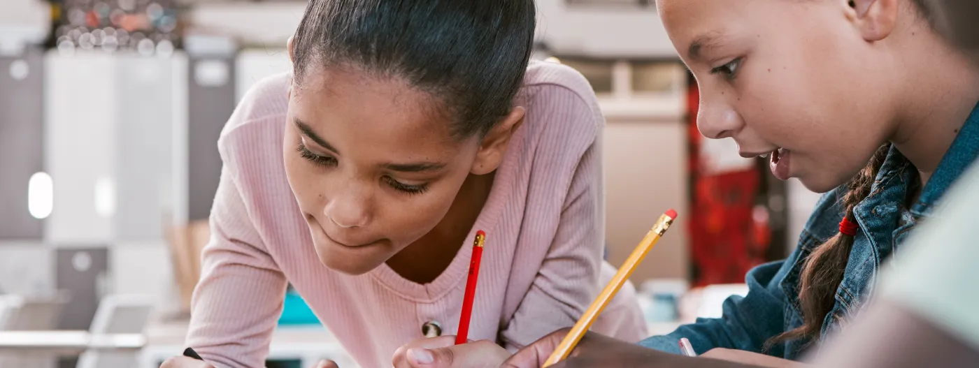 Tres niñas trabajan juntas en un salón de clases, escribiendo con lápiz en un cuaderno mientras comparten ideas y se concentran.