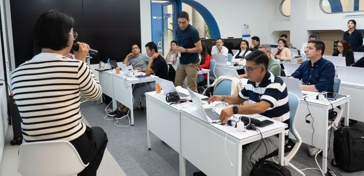 Foto de adultos en clase con cumputadores y docente mujer con micrófono