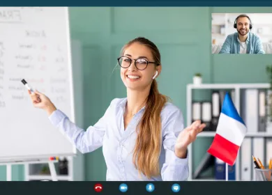 Profesora joven señalando tablero con palabras en francés, en curso virtual con alumno adulto en pantalla. Bandera de Francia