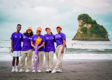 Foto de 5 estudiantes mujeres sonriendo con la antorcha olímpica, en la playa y en el fondo el mar y una gran roca