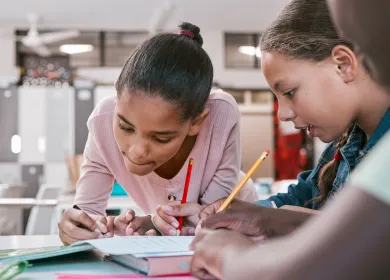 Tres niñas trabajan juntas en un salón de clases, escribiendo con lápiz en un cuaderno mientras comparten ideas y se concentran.
