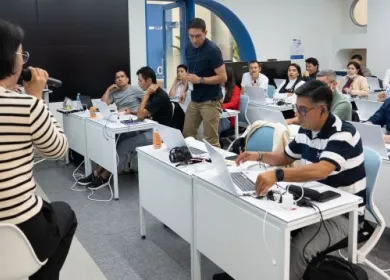 Foto de adultos en clase con cumputadores y docente mujer con micrófono